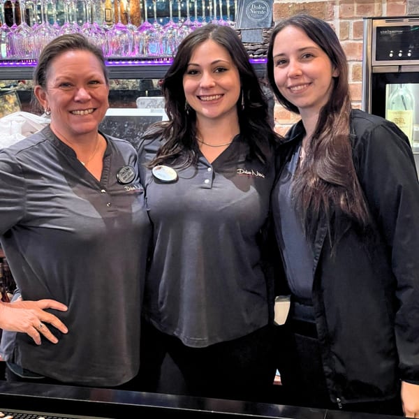 Three smiling women in uniform stand behind a bar with wine glasses and brick wall.