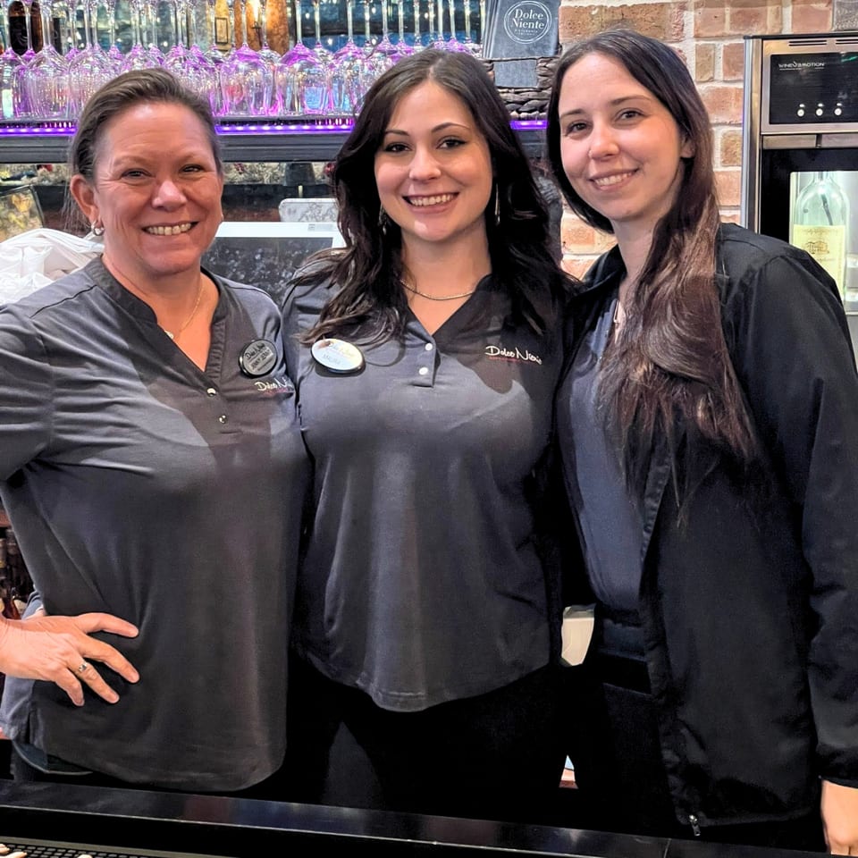 Three smiling women in uniform stand behind a bar with wine glasses and brick wall.