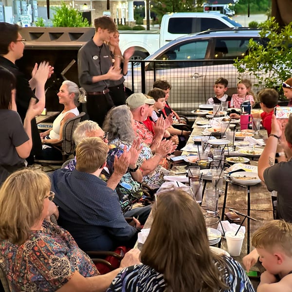 Restaurant staff and patrons, including children, clapping at a long outdoor dining table at dusk.
