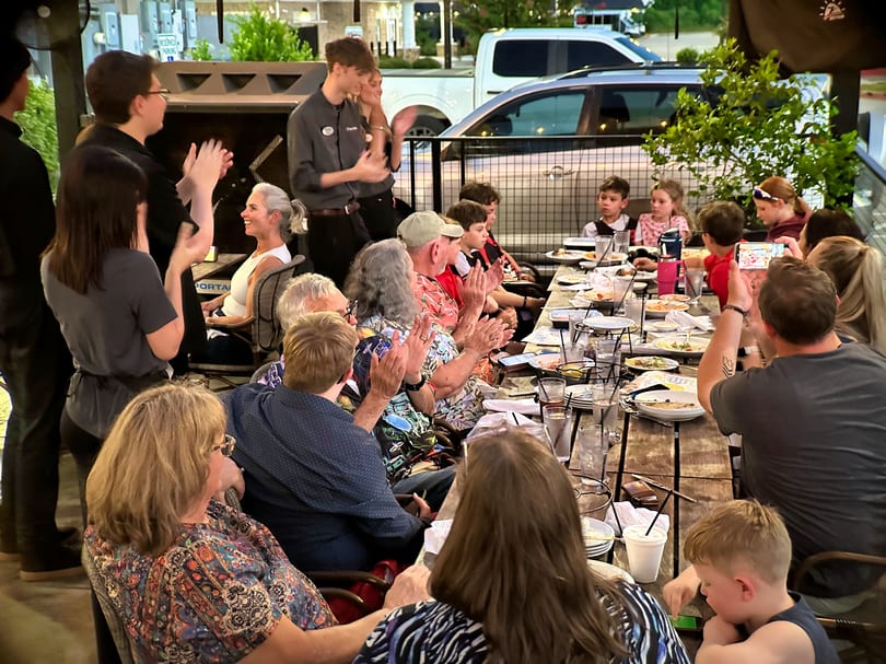 Restaurant staff and patrons, including children, clapping at a long outdoor dining table at dusk.