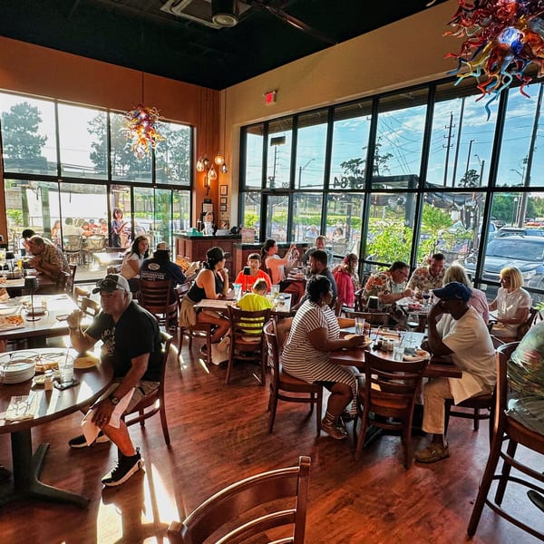 A busy restaurant dining room with many patrons seated at tables, bathed in natural light from large windows.