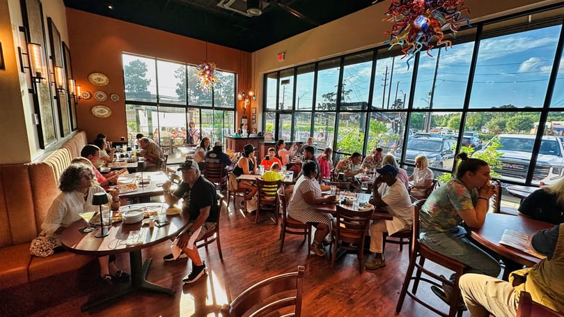 A busy restaurant dining room with many patrons seated at tables, bathed in natural light from large windows.